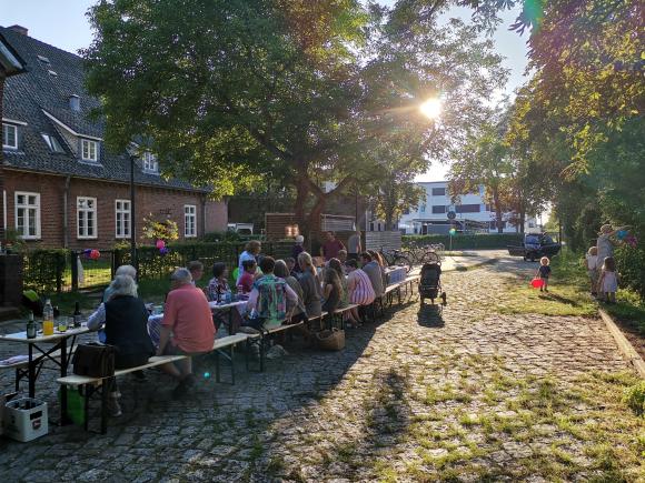 Nachbar:innen sitzen zusammen an einer langen Tafel vor einem alten Kasernengebäude. Die Sonne scheint auf die Szenerie. Ein kleines Kind steht neben den Bänken im Sonnenschein mit einem Luftballon