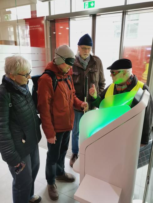 Eine Person mit Sehbehinderungs-Simulationsbrille steht vor einem Geldautomat